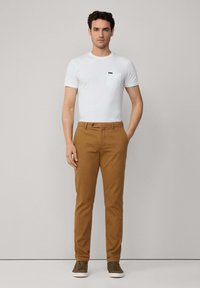 Camiseta blanca de manga corta con bolsillo en el pecho, combinada con pantalones chinos marrones. El modelo lleva zapatillas de color oliva. Diseño simple, colores neutros.