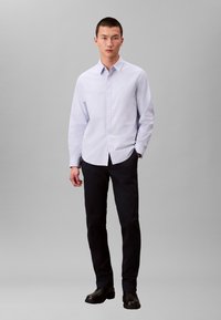 Chemise bleu clair à col boutonné, portée avec un pantalon foncé et des chaussures noires. La chemise a des manches longues et une coupe droite.