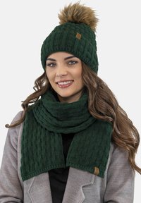 Gorro de lana verde con un pompón de pelo sintético y bufanda a juego con textura acanalada. Ambos artículos tienen un acento de logo de piel.