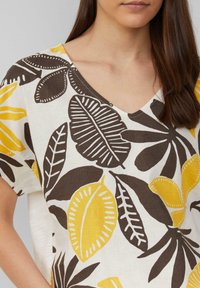 Korte mouw top van een lichte stof met een V-hals. Voorzien van een gedurfde botanische print in geel en bruin op een crèmekleurige achtergrond.