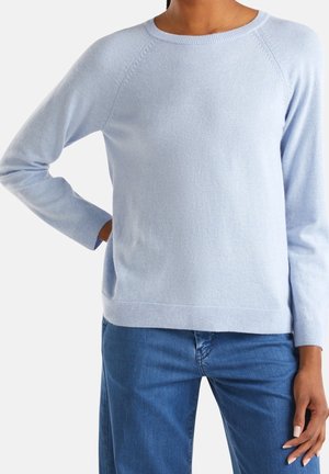 Personne portant un pull bleu clair à manches longues et un jean bleu, une main sur la hanche et l'autre détendue sur le côté.