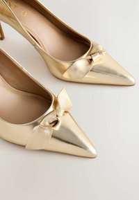 Zeltaini norādīti augstpapēžu apavi ar spīdīgu metāla apdari, ar loku akcentu uz priekšpuses un bēšas krāsas iekšējo zoli.