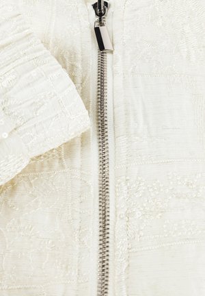 Close-up van wit stof met ingewikkelde bloemrijke borduurwerk en een centrale metalen rits met een rechthoekige trekker.