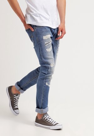 Jeans in denim azzurro chiaro con dettagli sbiaditi e strappi, abbinati a sneakers grigie basse con suole e lacci bianchi.