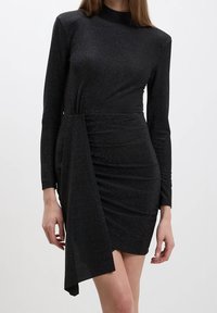 Kvinna klädd i en figurnära svart långärmad mini-klänning med asymmetrisk draperad tygdetalj framtill och en subtil glans.