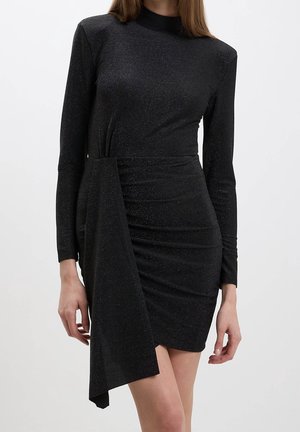 Woman wearing a fitted black long-sleeve mini dress with asymmetrical draped fabric detail on the front and a subtle shimmer.