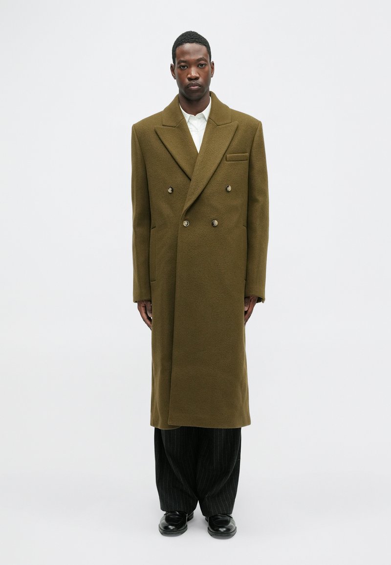 Vīrietis olīvu zaļā divrindu garajā mētelī, baltā kreklā, melnās svītrainās biksēs un melnās kurpēs, stāv uz viendabīgas baltas fona.