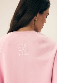 Różowa bluza z okrągłym dekoltem, z wytłoczonym białym tekstem na plecach: "BĄDŹ SOBĄ blovii." Miękka tkanina i luźny krój.