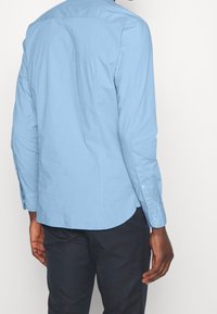 Man som bär en ljusblå långärmad skjorta med knappar och mörka byxor, sedd från ryggen mot en neutral bakgrund.