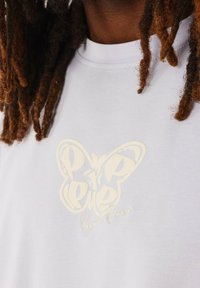 Camiseta blanca de algodón con un gran logo de mariposa en relieve de color crema suave y el texto "Project Paris" debajo.