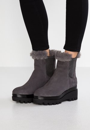 Femme portant des bottines en daim gris avec une bordure en fourrure et des semelles épaisses noires, associées à un pantalon noir moulant.
