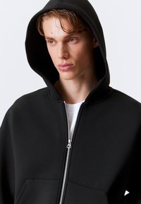 Sort zip-up hoodie med fleece tekstur, der har en stor hætte, sølvlynlås og kængurulomme. Bæres over en hvid T-shirt.