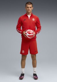 En atlet i en rød træningsdragt holder en rød og hvid fodbold, iført sorte støvler og hvide sokker, mod en grå baggrund.