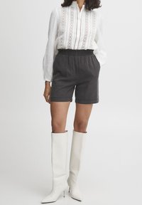 White blouse with textured fabric and lace embellishments, paired with black shorts and tall white heeled boots, standing on a neutral background.