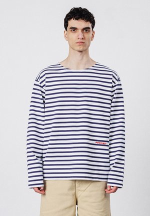 Młody mężczyzna z krótkimi, kręconymi, ciemnymi włosami, ubrany w białą koszulę z długim rękawem z ciemnymi poziomymi paskami oraz beżowe spodnie, stojący na jednolitym tle.