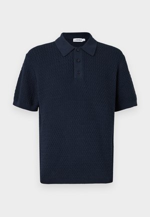 Mann steht mit einer Hand in der Tasche, trägt ein navy strukturiertes kurzärmeliges Polo, braune Hose und weiße Sneakers auf einfarbigem Hintergrund.