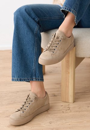 Jambes croisées, portant un jean bleu et des baskets en daim beige, assis sur une chaise en bois clair et tissu sur un sol en bois.