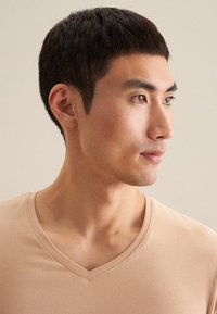 T-shirt beige en V fabriqué en tissu doux, avec une coupe ajustée et des manches courtes, présenté sur le cou et l'épaule d'un modèle masculin.