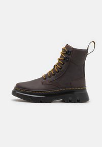 Botín de cuero marrón con cordones amarillos, elementos de hardware negro, cuello acolchado y una suela de goma negra robusta. Presenta detalles de costura amarilla.