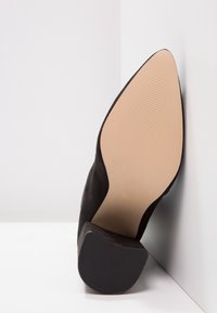 Chaussure à talon noir en daim doux avec un bout pointu, une semelle extérieure noire texturée et une semelle intérieure beige lisse. Design minimaliste.