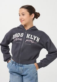 Szara bluza z kapturem na zamek błyskawiczny z białym i różowym napisem "BROOKLYN". Posiada kaptur na sznurki oraz ściągacze przy mankietach, w połączeniu z jasnoniebieskimi spodniami dżinsowymi.