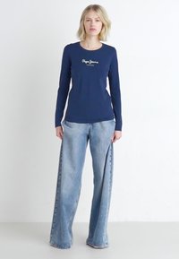 Marineblauwe langesleeve shirt met logo-detail aan de voorkant, gecombineerd met lichtblauwe wide-leg jeans met rafelzoom en subtiele vervaging.