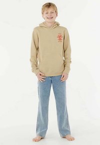 Beżowa bluza z kapturem i przednią kieszenią oraz czerwonym logo, zestawiona z jasnoniebieskimi szerokimi spodniami dżinsowymi. Modelka stoi boso.