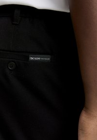 Prim-plan al unui buzunar de pantaloni negri cu un buton negru și o etichetă inscripționată "DENIM TOM TAILOR" lângă brațul unei persoane.