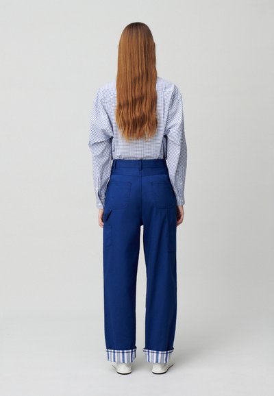 Pantaloni cargo blu con tasche laterali, a girocollo alle caviglie e con una fantasia a scacchi. Abbinati a una camicia a maniche lunghe con motivo a scacchi.