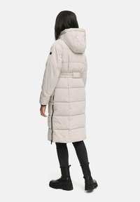 Lichtgrijze pufferjas met een capuchon, een ceintuur in de taille, een gewatteerd patroon en zijritsen, gecombineerd met zwarte slim-fit broeken en chunky laarzen.