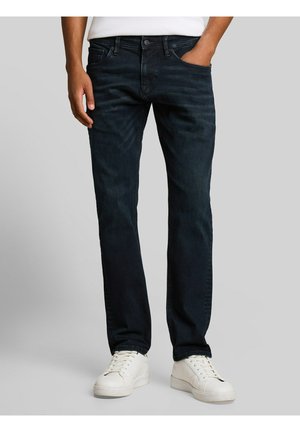 Mann trägt dunkelblaue Jeans und weiße Sneaker, steht mit einer Hand in der Tasche vor einem schlichten Hintergrund.