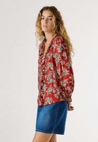 Blouse florale rouge avec un col froncé, des manches longues et une coupe ample, associée à une jupe en jean bleue. Le motif présente de grandes fleurs crème.