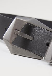 Ceinture en cuir noir avec une boucle rectangulaire en métal argenté. La boucle présente un design géométrique unique et une épingle en métal coulissante pour le fermeture.