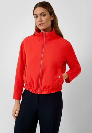 Femme portant une veste rouge vif à fermeture éclair avec un col montant et des poches avant, associée à un pantalon noir, debout devant un fond uni.