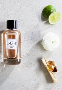 Le parfum Karl Lagerfeld Tokyo dans un flacon en verre transparent avec un bouchon noir, accompagné de moitiés de lime, d'une fleur blanche et de pierres d'ambre.