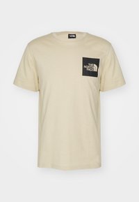 Camiseta de algodón beige con un bolsillo cuadrado negro que presenta texto de logo en blanco. Cuello redondo clásico y mangas cortas, con una textura suave.