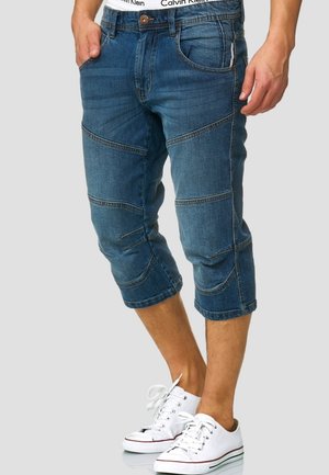 Homme portant un short en jean bleu jusqu'aux genoux, ceinture blanche de sous-vêtement Calvin Klein visible et baskets basses blanches, debout devant un fond uni.