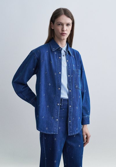 Femme portant une veste en denim bleu foncé assortie à un pantalon avec de petits motifs à pois blancs, chemise à col bleu clair, debout devant un fond uni.