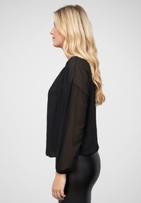 Blusa nera con maniche lunghe trasparenti e una vestibilità ampia. Presenta un collo rotondo e polsi elasticizzati. Indossata con leggings neri a vita alta.