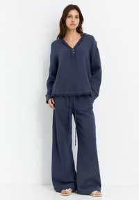Navy blauwe, loszittende loungeoutfit gemaakt van zachte stof. De top heeft een v-hals en knoopdetails, met rafelige randen. Broeken met wijde pijpen.
