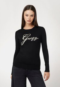 Maglione nero realizzato in tessuto morbido, con maniche lunghe e orlo a coste. La parte anteriore presenta un design del logo in strass argentati.