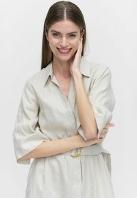 Światło beżowa sukienka lniana z kołnierzykiem, rękawami trzyczwartowymi, talią z paskiem i miękką fakturą. Modelka uśmiecha się, pozując na białym tle.