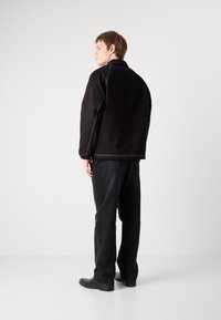 Giubbotto bomber nero con dettagli di cucitura bianca, vestibilità rilassata, tasche laterali con zip, indossato con pantaloni neri larghi e stivali neri.