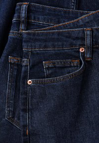 Gros plan d’un jean en denim bleu foncé montrant les poches avant, les passants de ceinture et les détails de couture orange.