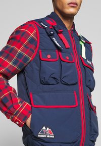 Marinfärgad quiltad väst med röda accenter, flera fickor med lappar och en logotyp-lapp. Bärs över en långärmad skjorta med röd och marinrutigt mönster.