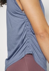 Närbild på en persons sida som visar en blå rynkad ärmlös topp och lavendelfärgade leggings mot en enfärgad bakgrund.