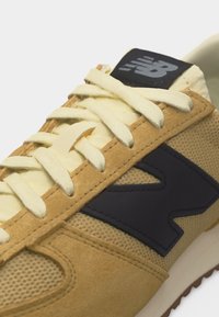 Ανοιχτό καφέ και μπεζ αθλητικό παπούτσι με εκρού κορδόνια και μαύρο λογότυπο "N" στη πλευρά, που δείχνει υφές από δίχτυ και σουέντ.