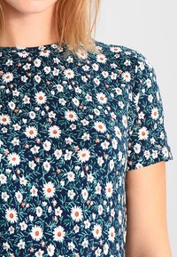 Top navy de manga corta, con un estampado floral de margaritas blancas y centros naranjas, sobre un fondo de hojas verdes.