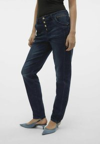 Jeans de mezclilla azul oscuro con corte recto, que presentan botones dorados y un acento decorativo en el bolsillo. Combinados con tacones azul claro.