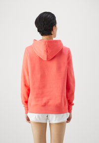 Felpa corallo con cappuccio regolabile e maniche lunghe. Realizzata in tessuto morbido, presenta orlo e polsini a coste. Indossata con pantaloni corti bianchi.